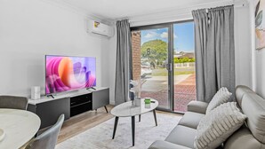 Ein modernes Wohnzimmer mit Flachbildfernseher, einem grauen Sofa, einem runden Couchtisch und einem Essbereich mit weißem Tisch und Stühlen.