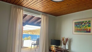 A hotel room with a bed, a desk with a TV, a painting on the wall, and a view of the sea through the curtains. - Art Sounion, Seafront Elegance (Sounion, Lavreotiki)