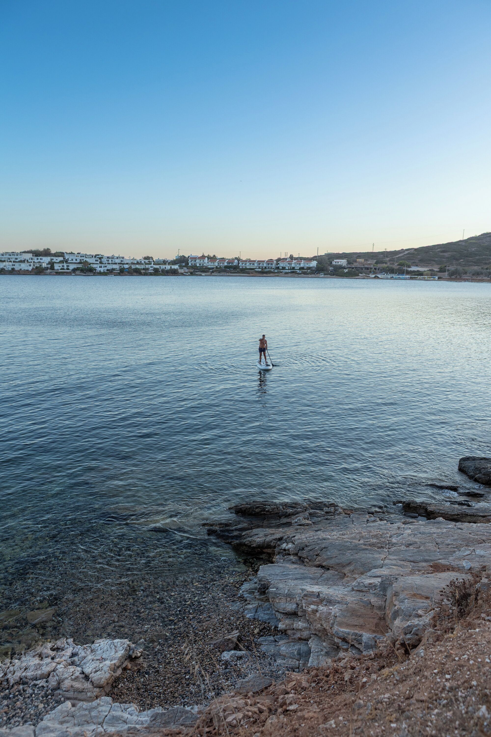 Eine Person steht auf einem Paddleboard auf ruhigem Gewässer, im Hintergrund eine felsige Küste und Gebäude.