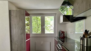 Et moderne køkken med indbygget ovn, mikroovn og bordplade i rustfrit stål. To vinduer giver naturligt lys og udsigt til grønne områder.