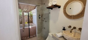 Une salle de bain dotée d’une douche à parois de verre, d’un lavabo avec un récipient en pierre et d’un miroir dont le cadre est fait de fibres tressées.