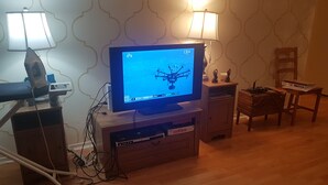 Ein Fernseher zeigt eine Drohne, eine Lampe, einen Stuhl und einen Holzschrank mit DVD-Player.