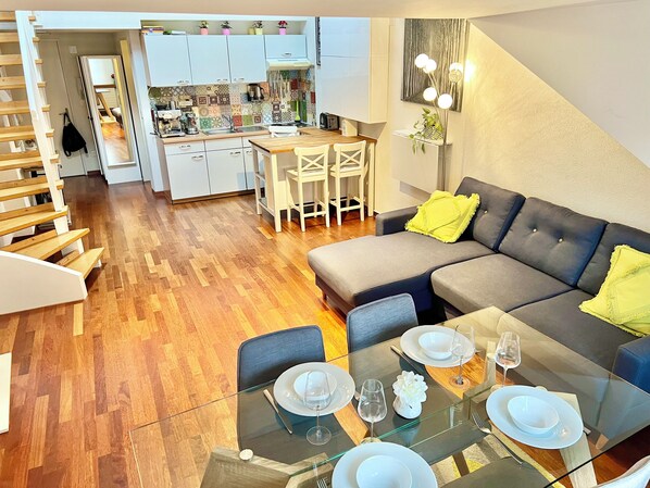 A modern living room with a glass dining table set for four, a grey sofa with yellow cushions, and a kitchen in the background. - Central Old Town Gem with Rare 40m² Terrace & River Views (Zürich)