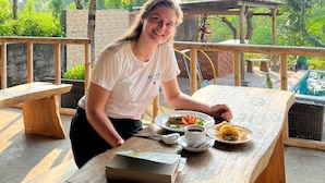 A person seated at an outdoor wooden table with a thatched roof overhead, surrounded by greenery and a pool in the background. - Eco Moringa Garden (Bali)