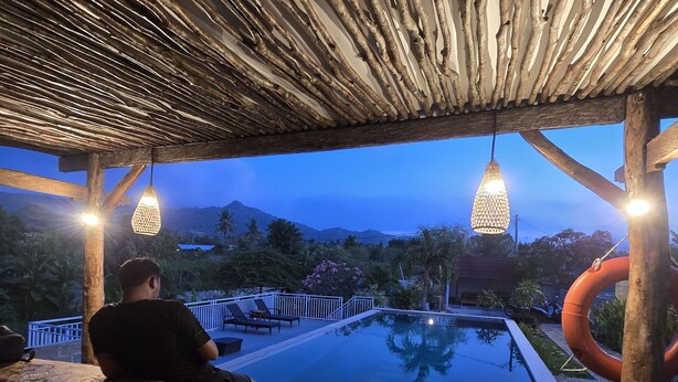 A wooden poolside area with a person seated at a table, a pool, and a view of the mountains. - Eco Moringa Garden (Bali)