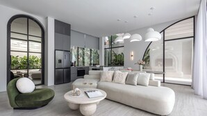 Una sala de estar moderna con sofá, sillón y mesa de centro, un área de cocina y ventanas grandes con vista a follaje.