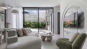 Una sala de estar moderna con sofá, sillón y mesa de centro. Se aprecia un ventanal con vistas a zonas verdes y una piscina.