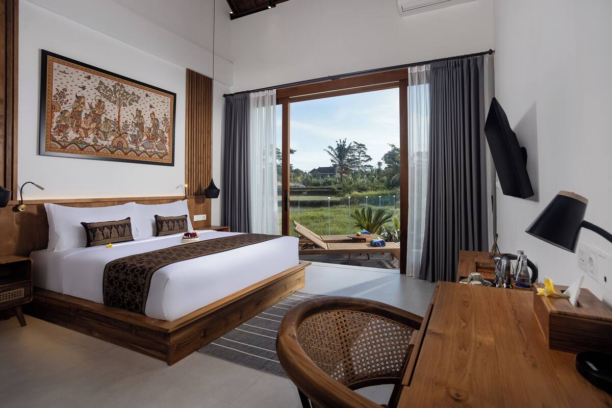 A hotel room with a large bed, a desk, a chair, and a view of a garden through the window.