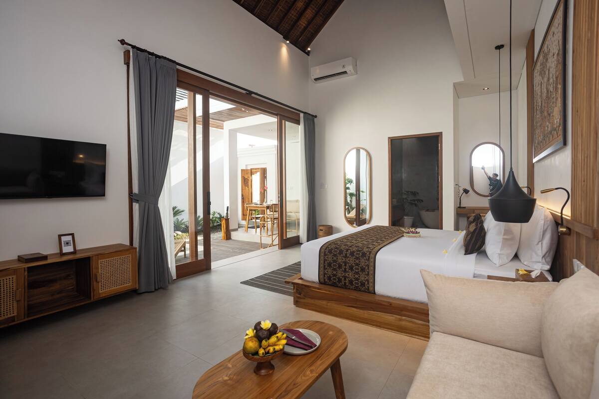 A modern hotel room with a bed, a sofa, a wooden table with fruit, a TV, and a sliding glass door to a balcony.