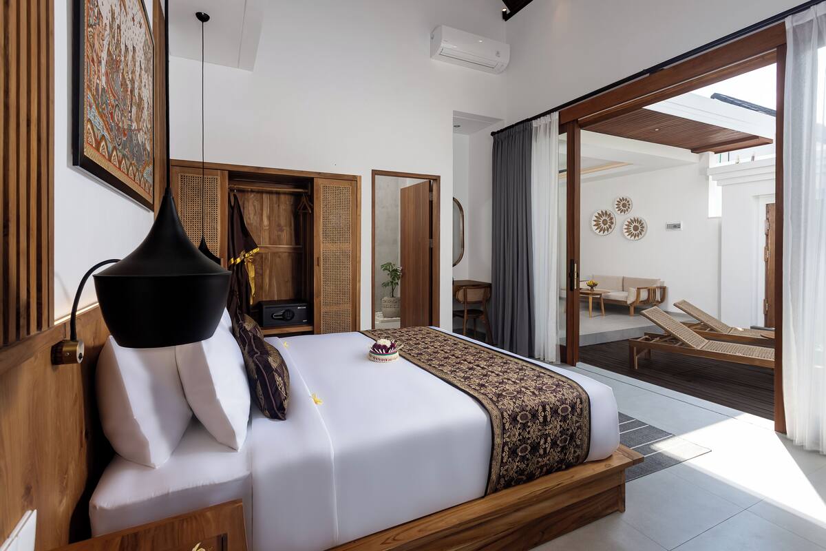 A bedroom with a bed, bedside table, lamp, and a view of an outdoor seating area.