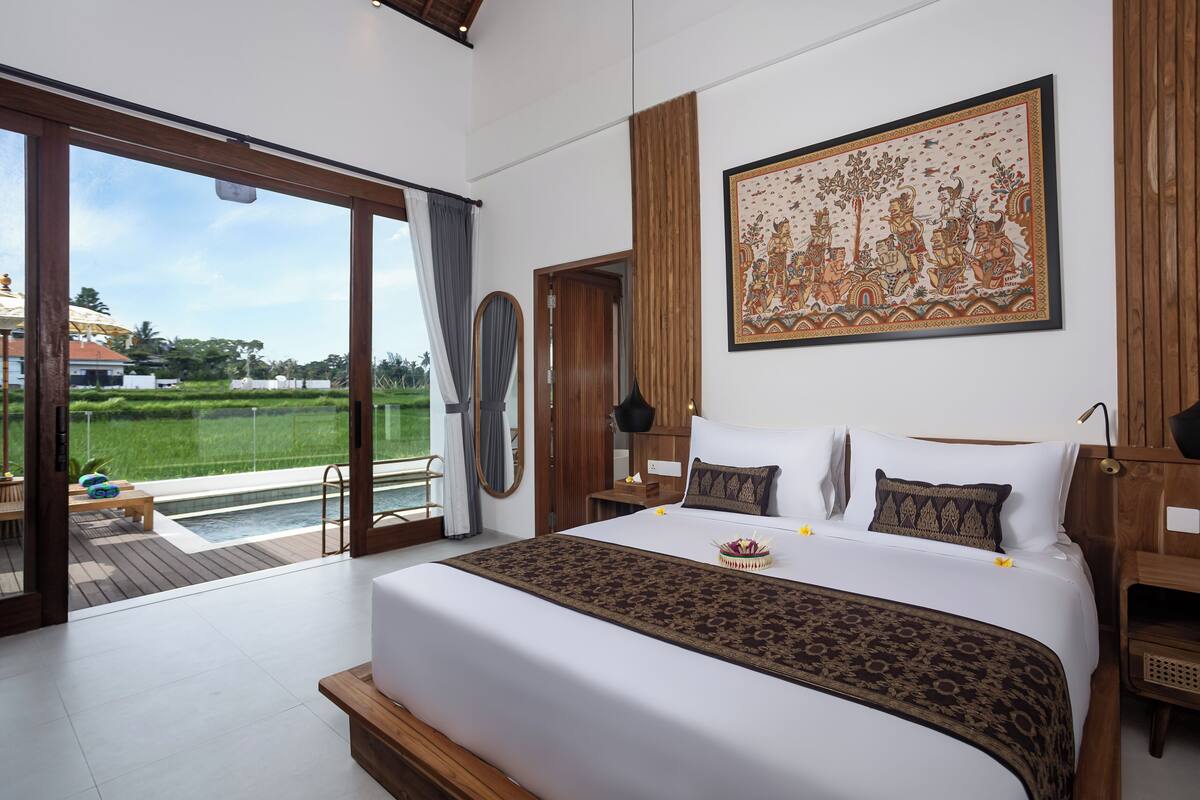A spacious bedroom with a large bed, a wooden headboard, a large window, and a view of a green landscape.