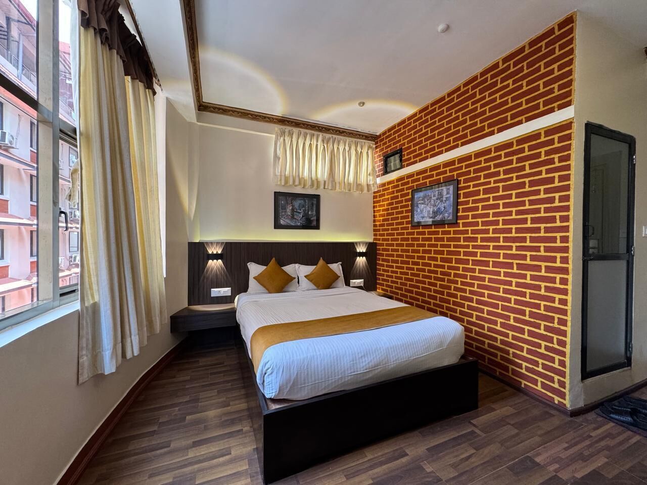 Una habitación de hotel con pared de ladrillo decorativa, una cama grande con cabecera, una mesita de noche y una ventana con cortinas.