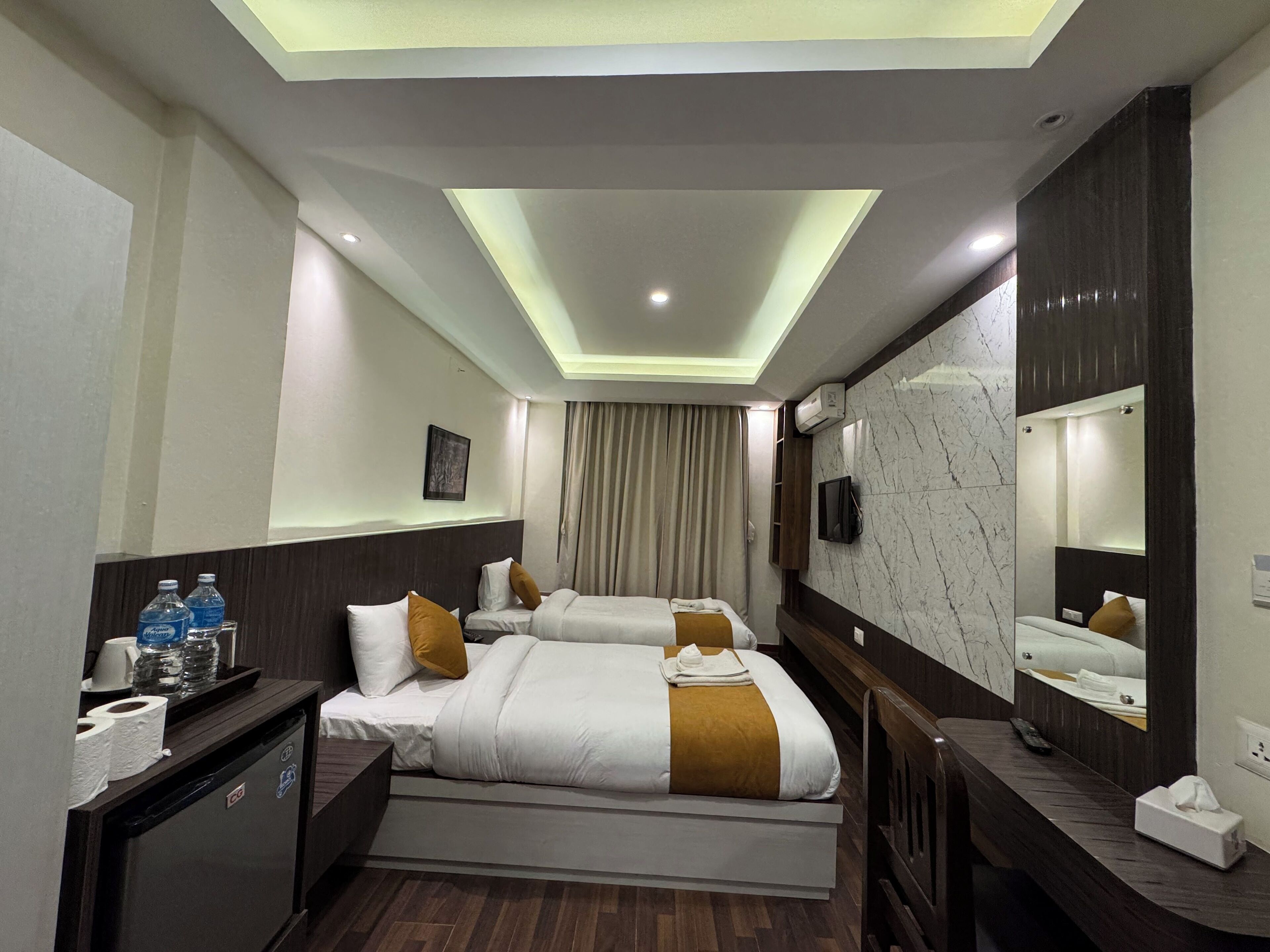 A hotel room with two beds, a minibar, a mirror, and a ceiling light.