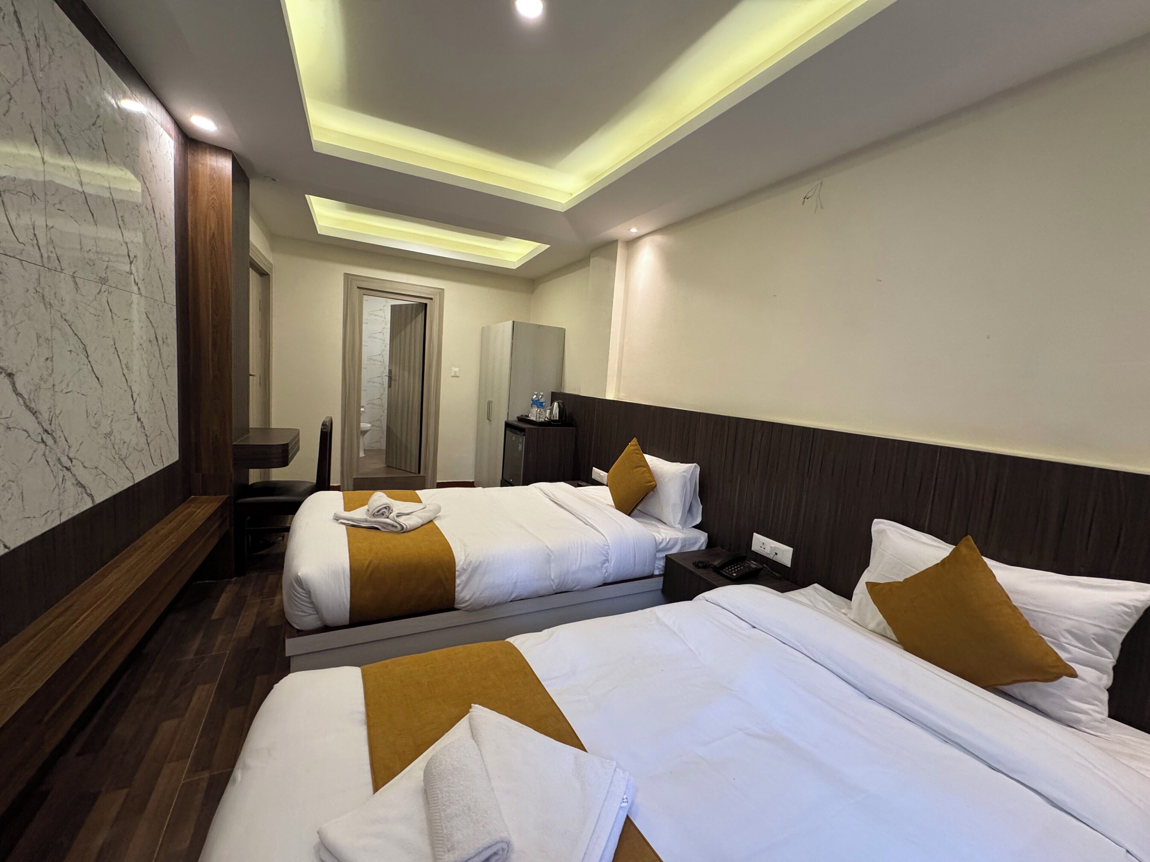 A hotel room with two beds, a wooden bench, a desk, a wardrobe, and a bathroom with a marble wall.