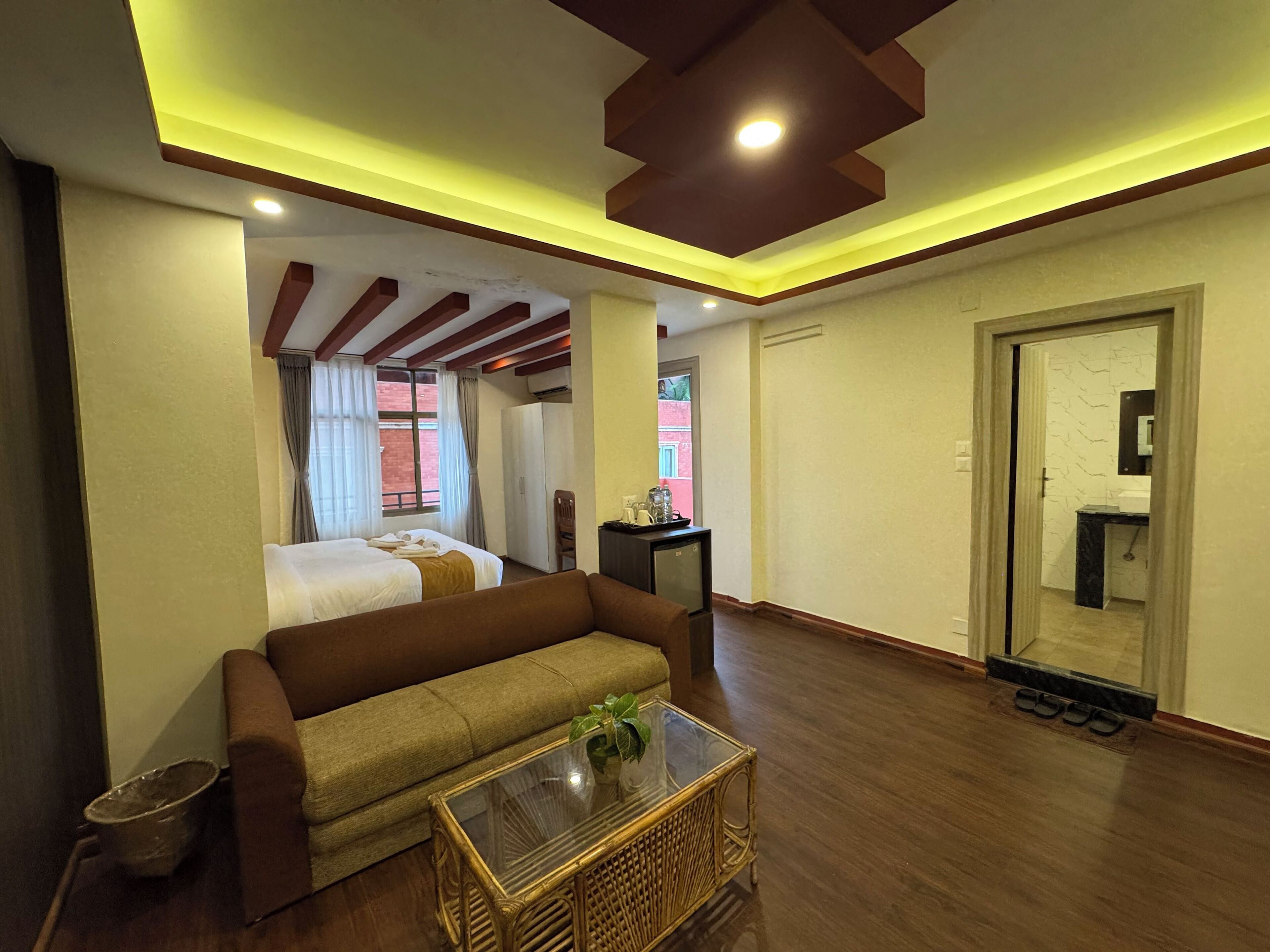 A hotel room with a bed, a sofa, a small table with a plant, a trash bin, and a door leading to a bathroom.