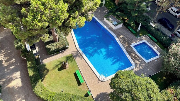 Una piscina rectangular con una piscina más pequeña contigua, rodeada de vegetación cuidada y una terraza pavimentada.