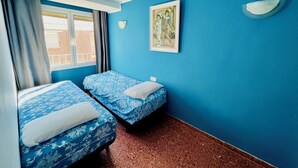 Una habitación con dos camas, una ventana con cortinas, una lámpara fijada a la pared y un cuadro enmarcado.