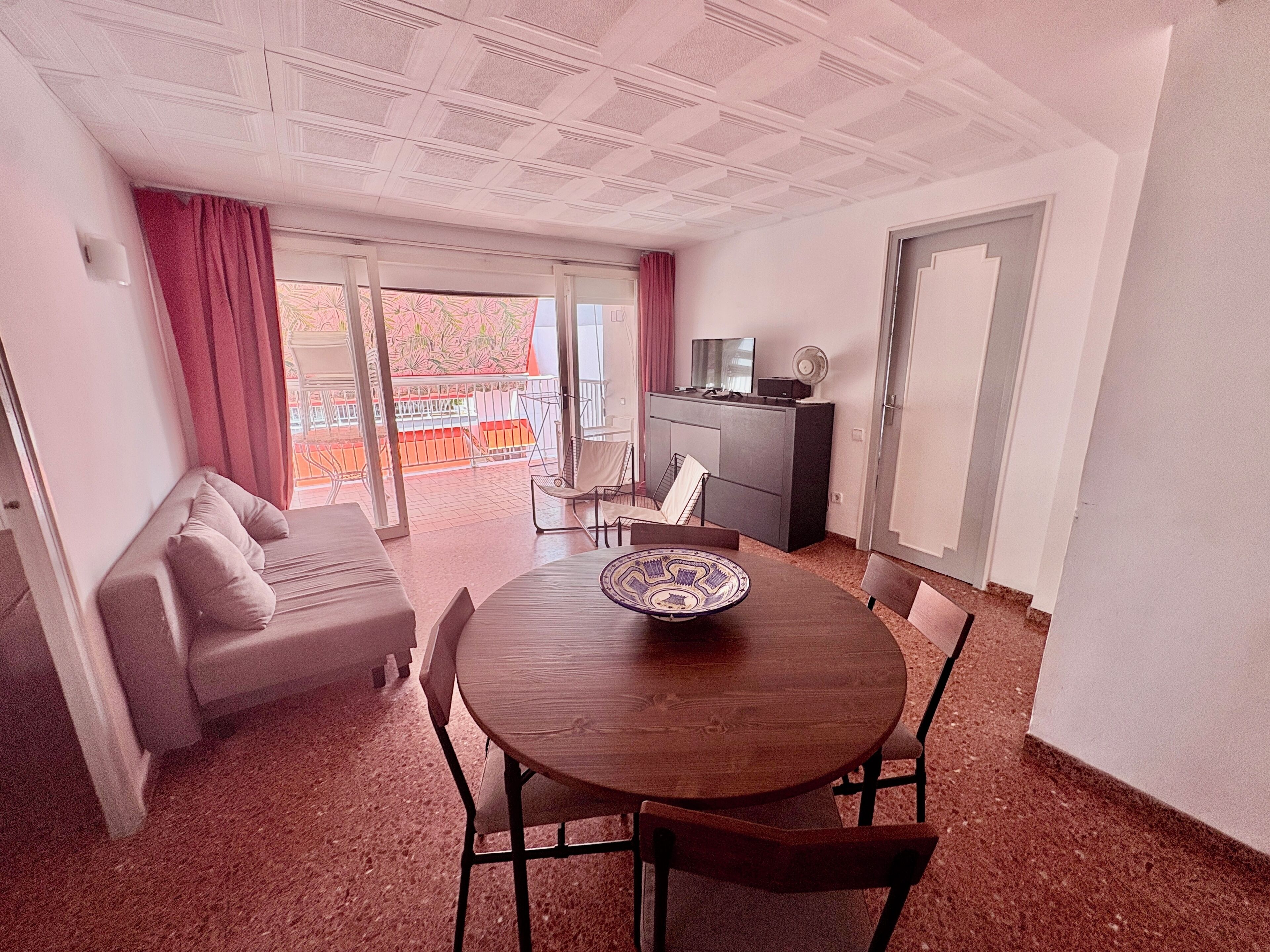 Una habitación con una mesa redonda de madera, un sofá, un televisor y un balcón con vistas.