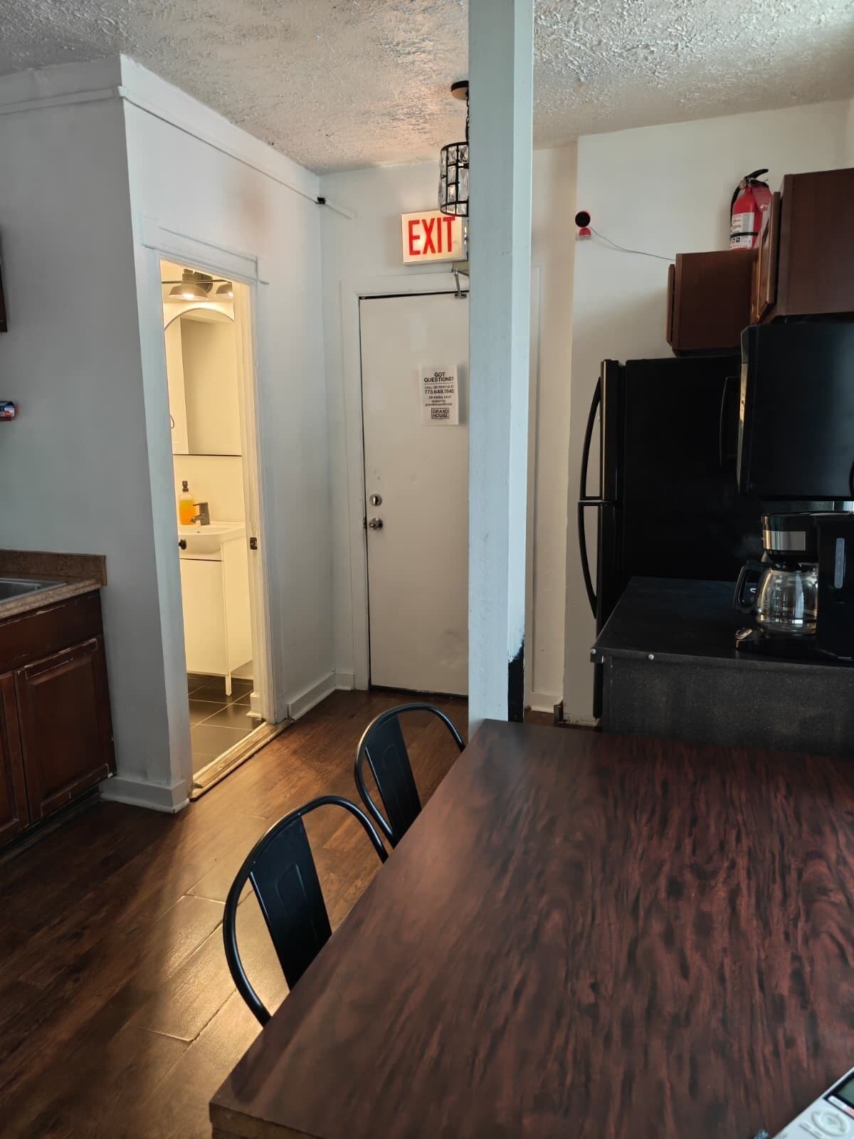 A kitchen with a dining table, chairs, a refrigerator, and an exit sign.