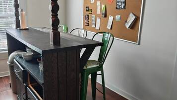 Una habitación con una mesa de madera, sillas, un tablón de anuncios y una ventana con persianas.