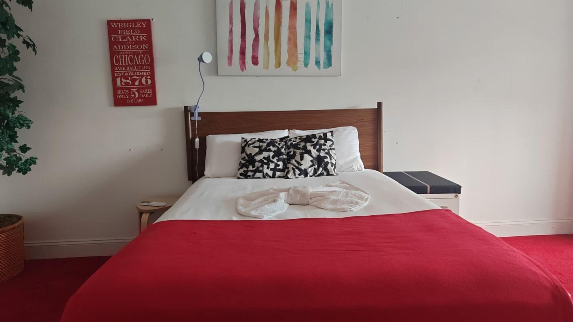 Habitación de hotel con colcha roja, cabecera de madera, mesita de noche con lámpara y cartel en la pared.