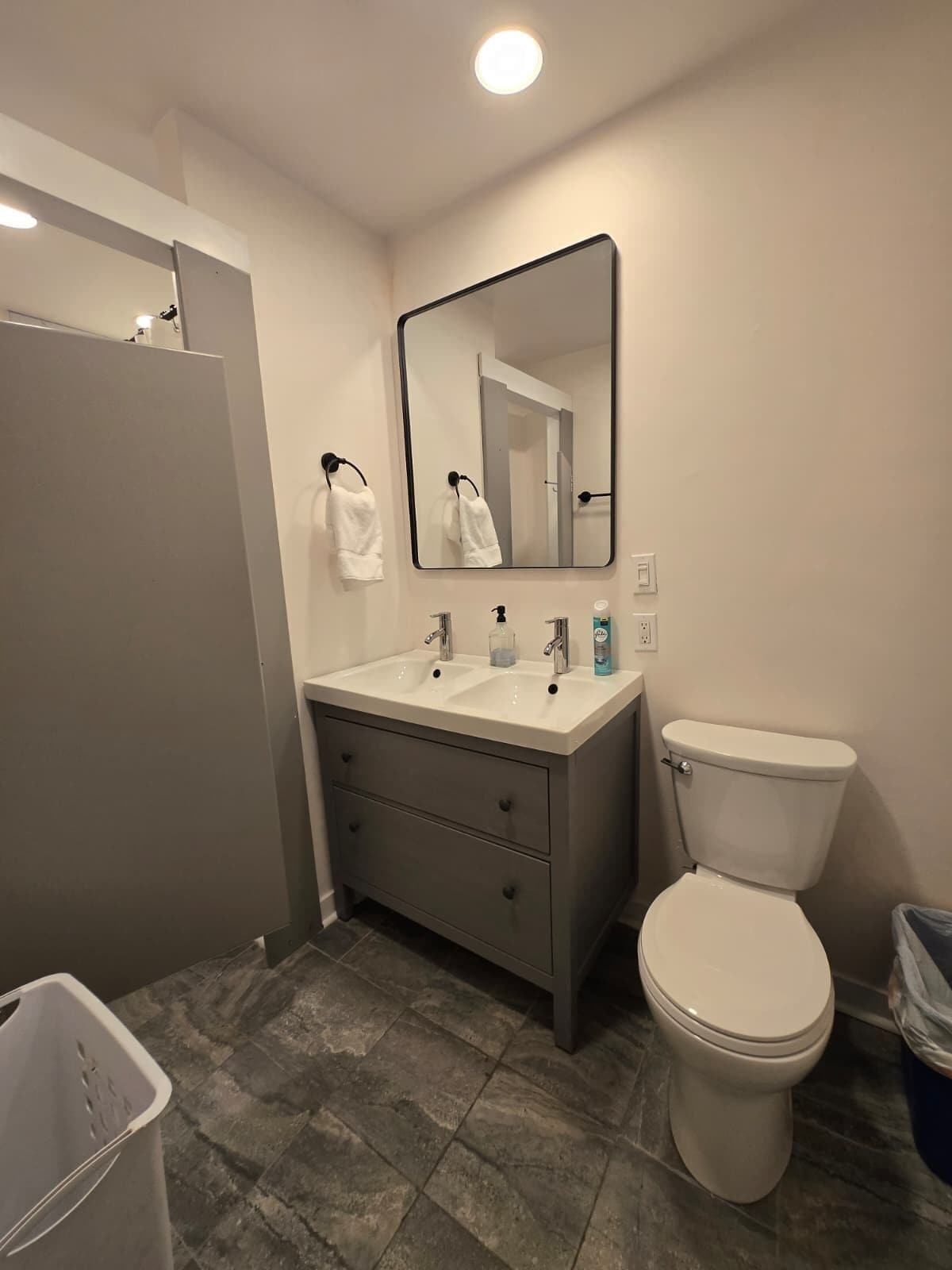A bathroom with a white sink, two faucets, a mirror, a toilet, and a trash bin.