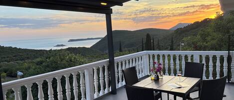 A balcony with a table and chairs, overlooking a coastal landscape with hills and a view of the sea.