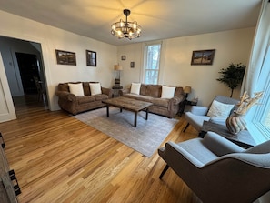 A living room with a wooden floor, two brown sofas, a coffee table, and a grey armchair. There is a chandelier, a window with a view, and a plant on a side table.