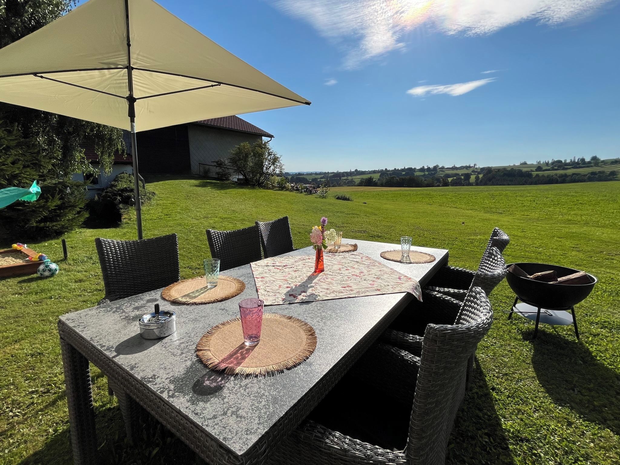 Un área de comedor al aire libre con una mesa preparada para seis personas, rodeada de sillas de mimbre, bajo un paraguas grande.