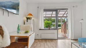 A modern, well-lit room with a wooden floor, a white sofa, a wicker chair, a small table with a lamp, and a view of a garden through a glass door.