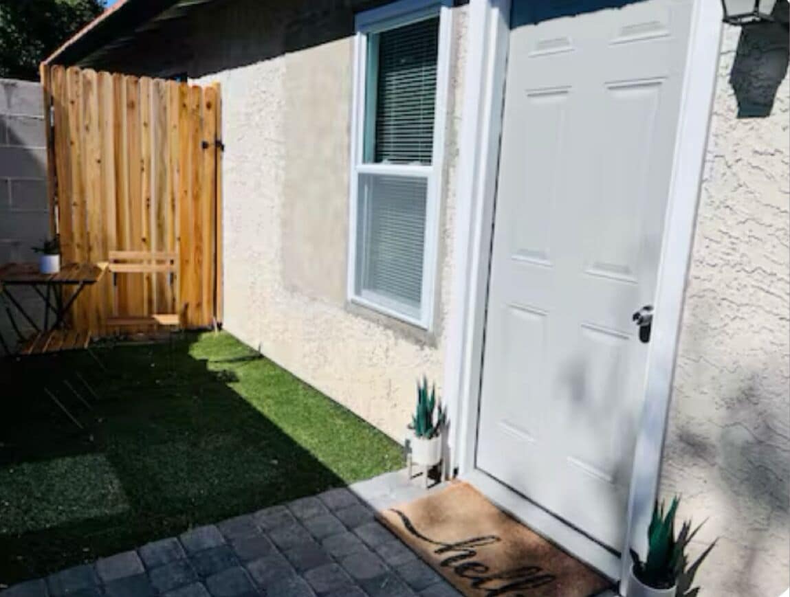 A house with a white door, a small window with blinds, a wooden fence, a patio with a table and chairs, and a welcome mat.