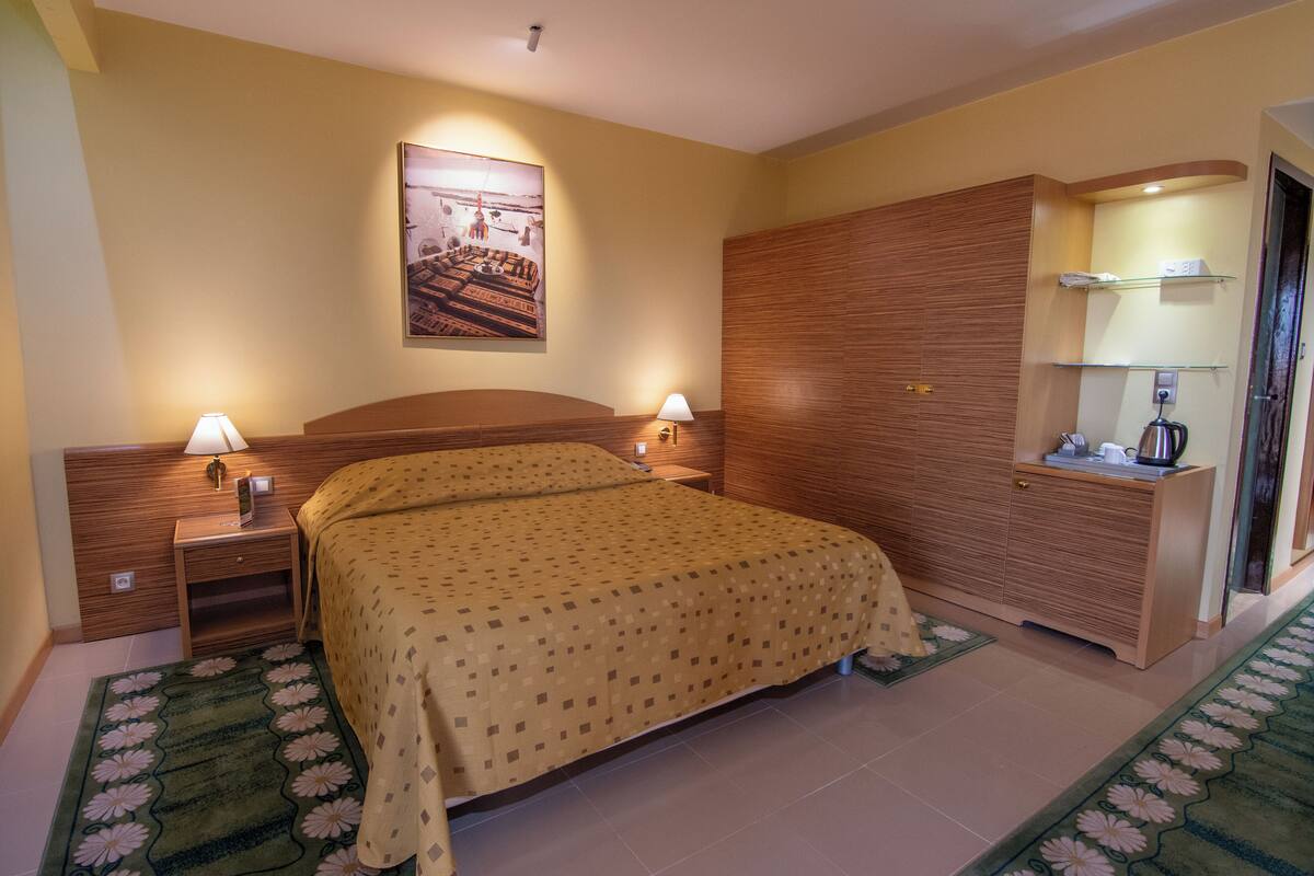 Een hotelkamer met een bed, nachtkastjes, lampen, een houten garderobe, een spiegel en een ingelijst schilderij aan de muur.
