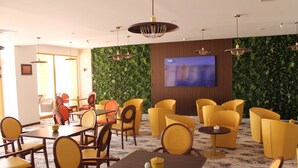 Een moderne hotellobby met geel en bruin zitmeubilair, een groot televisiescherm en een groene wand met planten.
