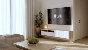 A modern living room with a flat-screen TV mounted on a white wall, a wooden coffee table, a beige sofa, and a large window with sheer curtains.