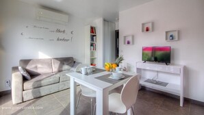 A modern living room with a sofa, a dining table with chairs, a bookshelf, and a flat-screen TV on a stand.