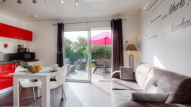 A modern kitchen with red cabinets, a dining area with a white table and chairs, a grey sofa, and a sliding glass door leading to an outdoor patio with a pink umbrella.
