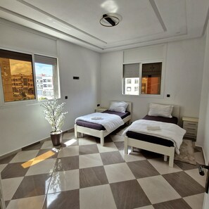 Habitación de hotel con dos camas, una planta en maceta y vistas a edificios a través de la ventana.