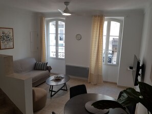 A living room with a sofa, a round coffee table, a dining table, and a TV. There are two windows with curtains, a ceiling fan, and a wall clock.