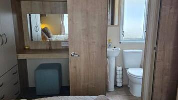 Un dormitorio con cama, un armario de madera, un taburete azulito y un baño con lavabo, inodoro y ventana.