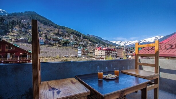 A wooden table and chair set on a balcony overlooking a mountainous village with buildings and snow-capped peaks. - Manali Heaven (Manali)