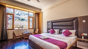 A hotel room with a large bed, a wooden headboard, a small table, and a chair. There is a window with a view of a mountainous landscape. - Manali Heaven (Manali)