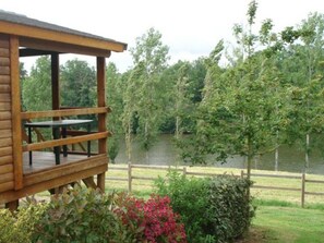 A wooden cabin with a deck overlooking a lake, surrounded by lush greenery and a wooden fence.