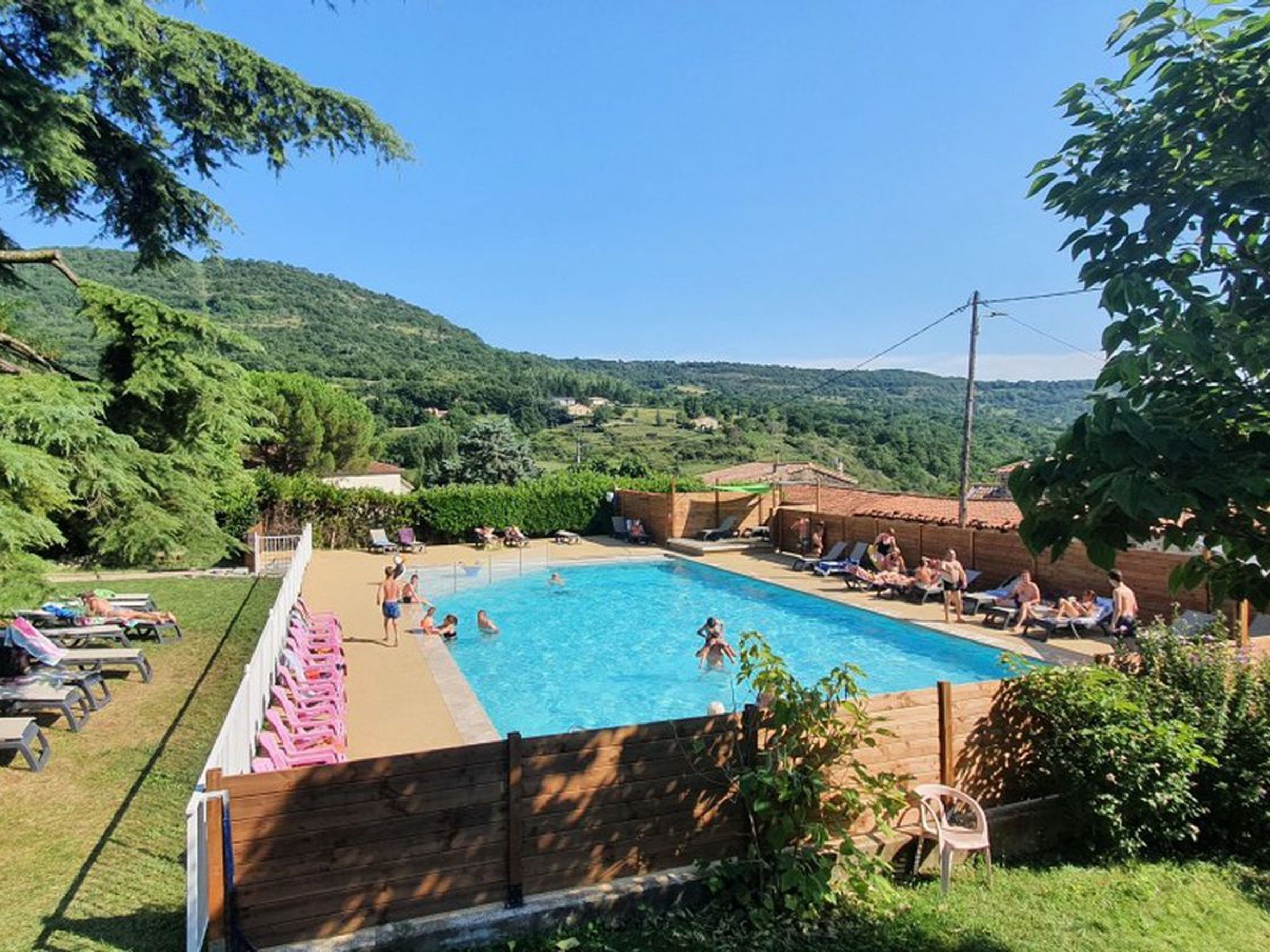 Une piscine entourée d’une clôture en bois, où l’on voit des gens nager et se détendre au bord de l’eau.