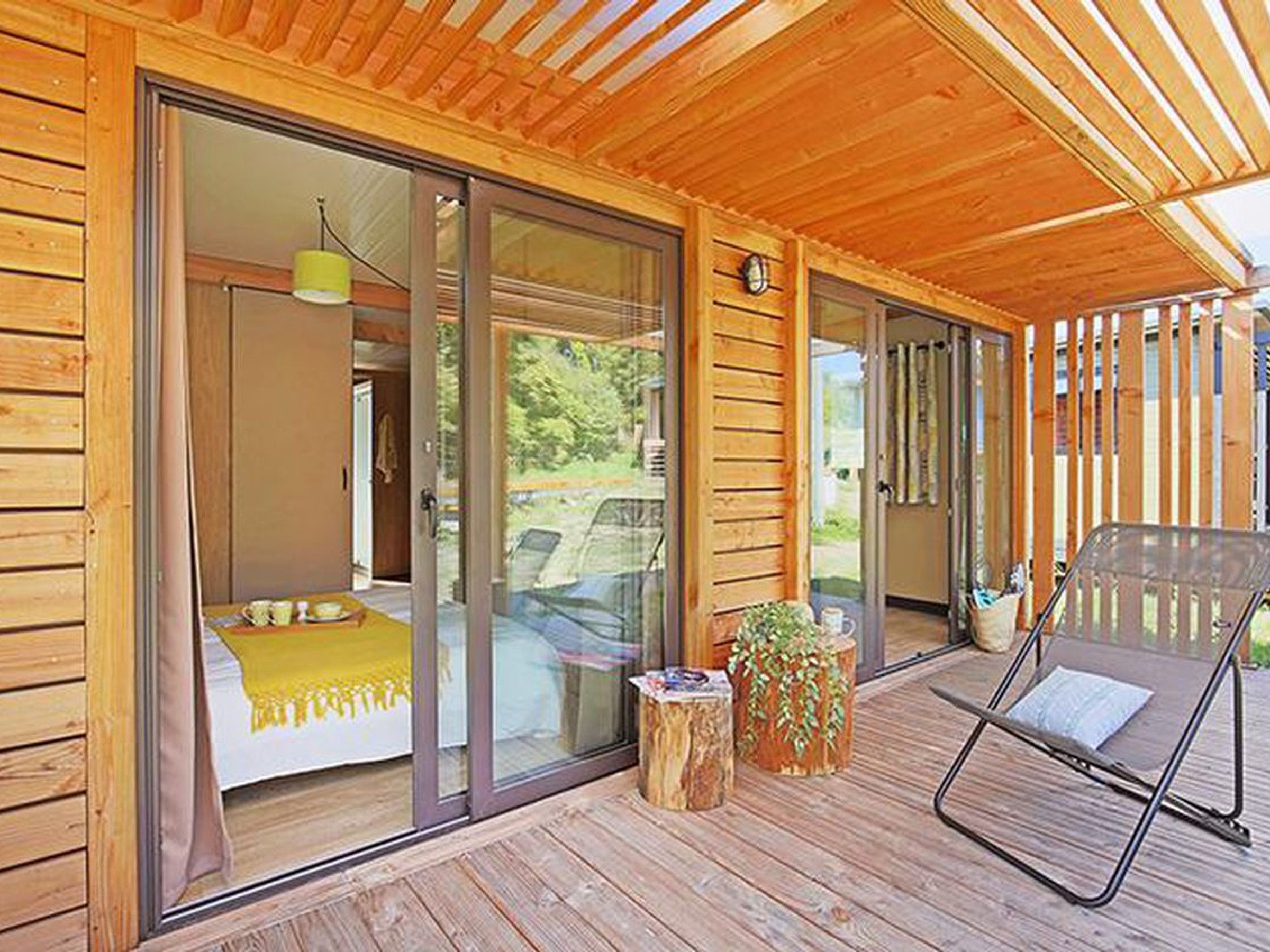Eine Holzhütte mit einer Schiebetür zu einem Schlafzimmer, das ein Bett und eine gelbe Decke zeigt. Vor der Tür befinden sich zwei Holzschemel mit Pflanzen.