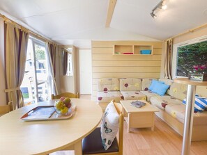 A cozy living room with a sofa, a dining table with fruit, and a window with curtains. - Mobile home with terrace and equipped kitchen (Pezuls)