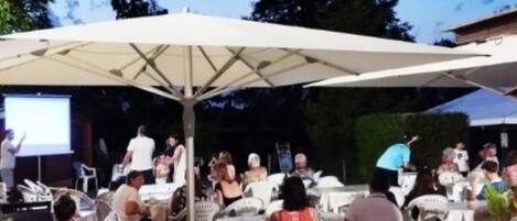 An outdoor event with tables and chairs under umbrellas, a stage with a presentation screen, and people gathered around.