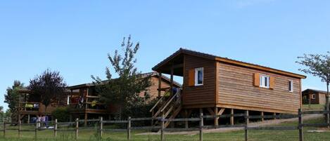 A wooden cabin with a balcony and a staircase leading up to it, surrounded by a wooden fence and greenery.