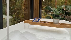 Une baignoire avec un plateau en bois sur lequel sont posés un livre et une tasse, le tout dans un décor offrant une vue sur la forêt.