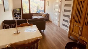 Ein Zimmer mit Esstisch, Stühlen, einer Couch, einem Fernseher und Blick durch große Fenster.