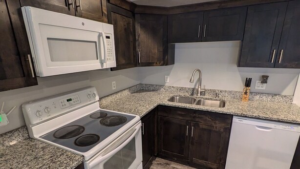 A kitchen with dark wood cabinets, a white microwave, a stove, a dishwasher, and a granite countertop. - Modern 1BR by Silicon Slopes – Workspace & 65” TV (American Fork)